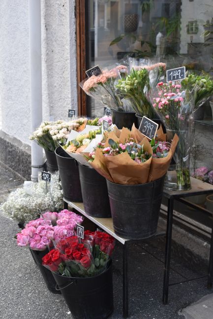 An outdoor display of fresh flower bouquets arranged in black buckets on a black metal table outside a flower shop. The arrangements feature a variety of seasonal blooms, including pink roses, white c