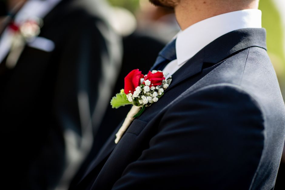Close-up of a groom dressed in a navy blue suit and white shirt, wearing a boutonniere consisting of a vibrant red rose, small white gypsophila flowers, and green foliage pinned to his lapel, against