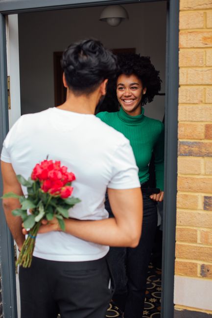 A woman presents a vibrant bouquet of red and pink roses with green foliage to another woman standing inside a doorway. The recipient, with curly black hair and a bright smile, wears a green turtlenec