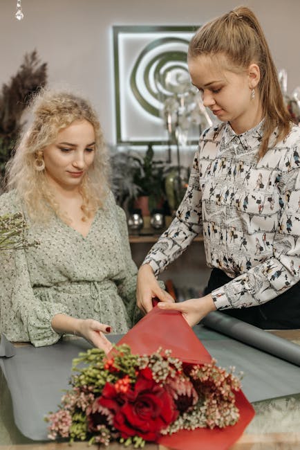 A professional florist, dressed in a patterned blouse, is wrapping a vibrant bouquet of red roses, pink carnations, and small white filler flowers in red floral tissue paper on a table. The arrangemen