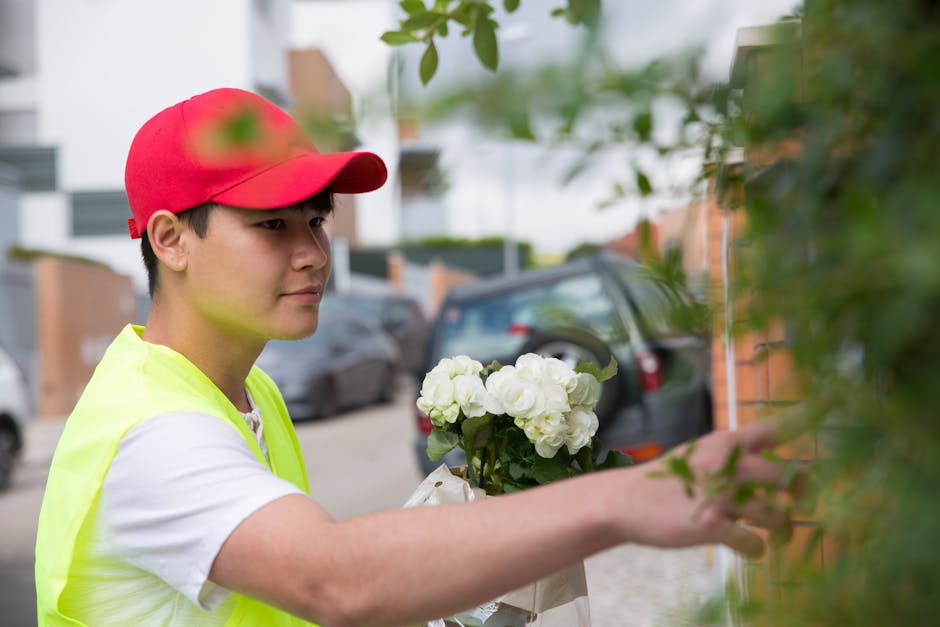 A young delivery person wearing a red cap and a high-visibility yellow vest is placing a floral gift outside a residential property, with a bouquet of fresh white hydrangeas wrapped in paper in hand. 